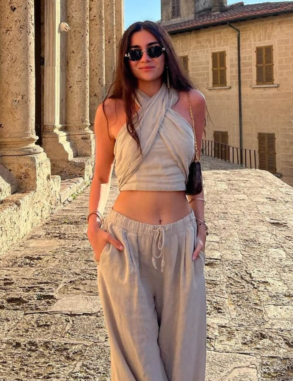 Modelo em cenário de rua antigo vestindo Conjunto Amara bege de casimira, com top cruzado no pescoço (frente única) e calça pantalona solta