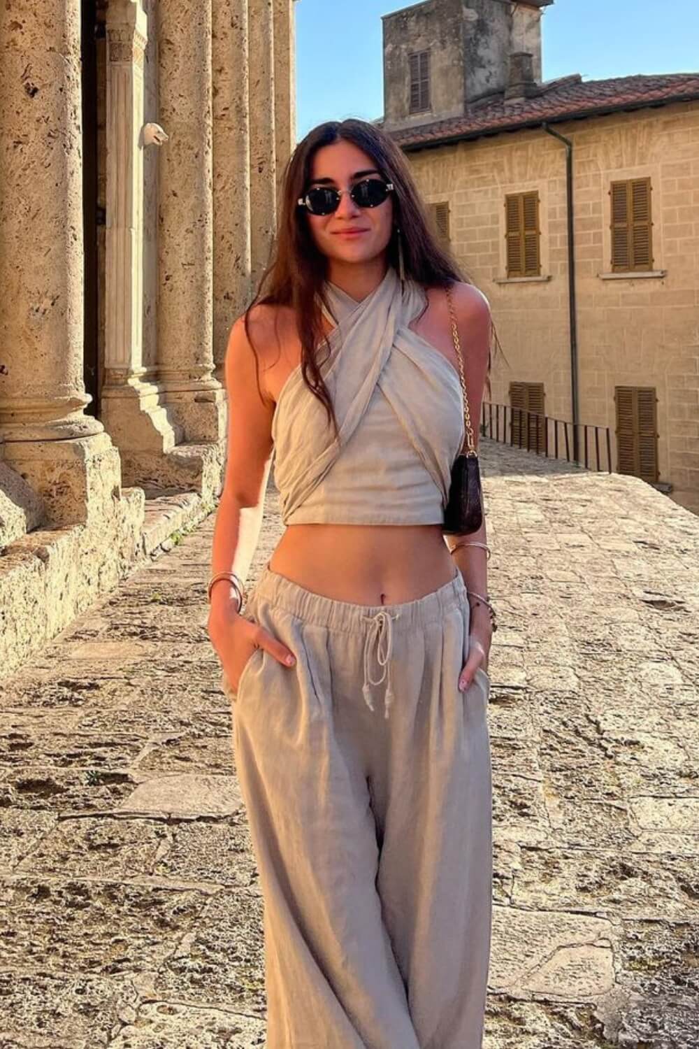 Modelo em cenário de rua antigo vestindo Conjunto Amara bege de casimira, com top cruzado no pescoço (frente única) e calça pantalona solta