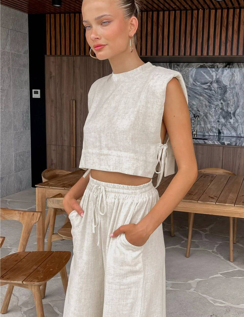 Modelo sentada vestindo Conjunto Giza de linho natural bege, composto por cropped com ombreiras e laços laterais, e calça pantalona solta