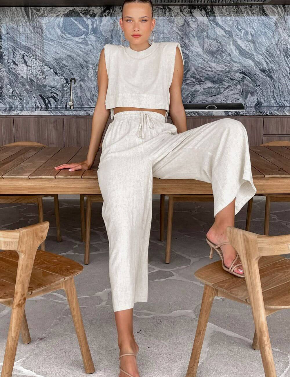 Modelo sentada vestindo Conjunto Giza de linho natural bege, composto por cropped com ombreiras e laços laterais, e calça pantalona solta