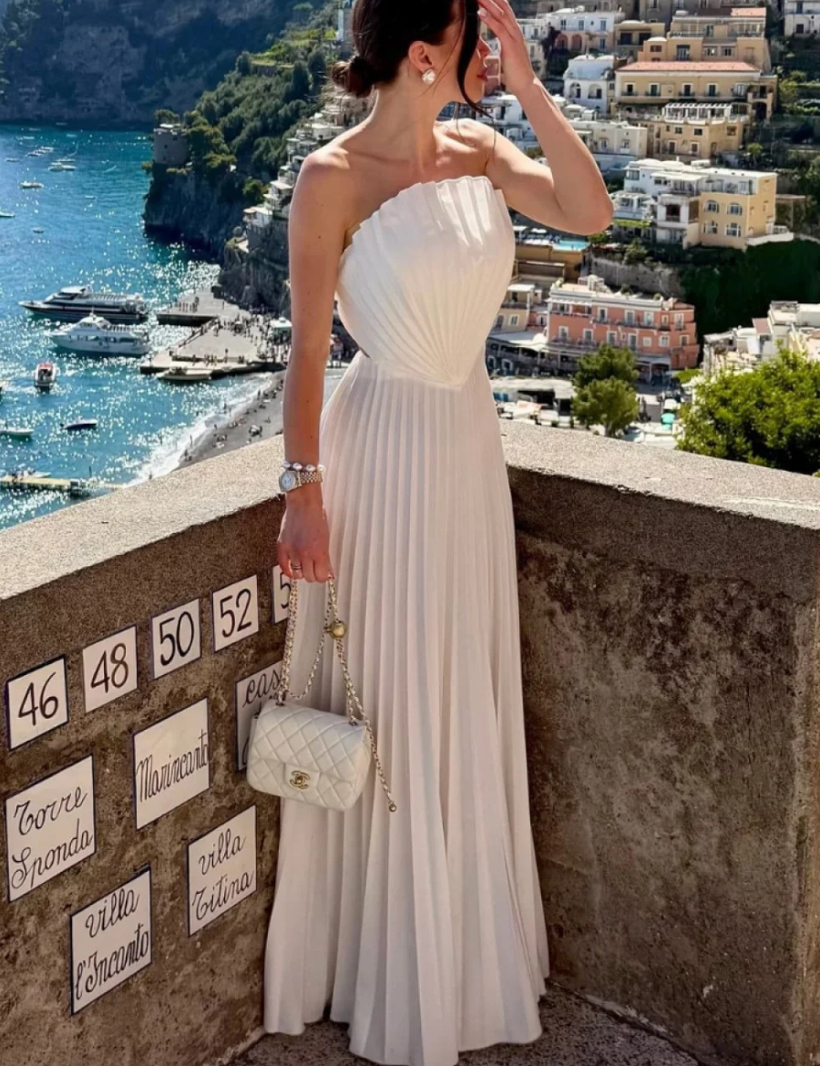 Modelo em cenário de Positano vestindo Vestido Keisy longo off white, todo plissado, com busto rígido em formato de concha e fenda lateral profunda.