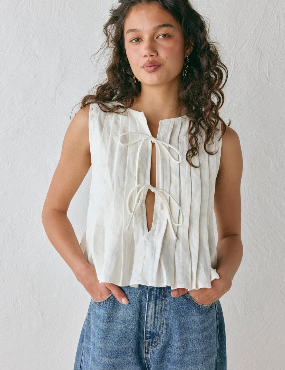 Modelo vestindo Top Bianca, uma regata de linho misto bege com textura plissada vertical, modelagem solta e fechamento frontal com dois laços delicados.