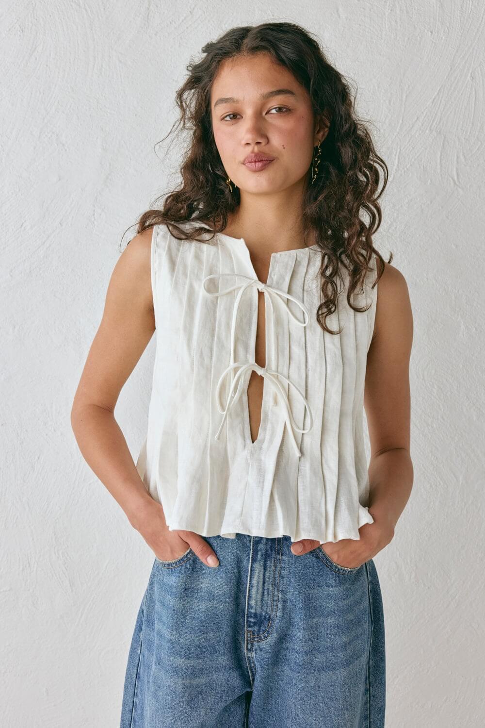 Modelo vestindo Top Bianca, uma regata de linho misto bege com textura plissada vertical, modelagem solta e fechamento frontal com dois laços delicados.